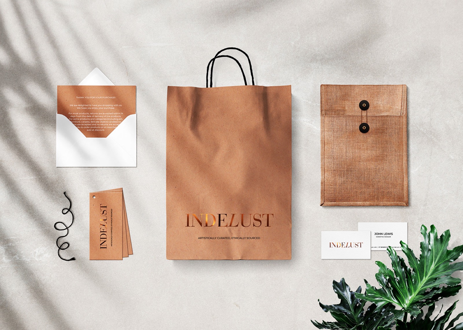 Indelust brand collateral flatlay: a kraft shopping bag with a copper-foil logo, hangtags, business cards, a jute envelope and a printed thank-you note, all in unbleached natural materials.